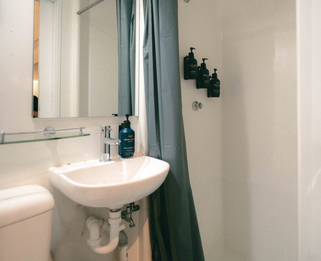 Small bathroom scene with white pedestal sink, exposed pipes beneath, and a blue bottle on the sink rim; shower curtain to the right and wall-mounted dark bottles.