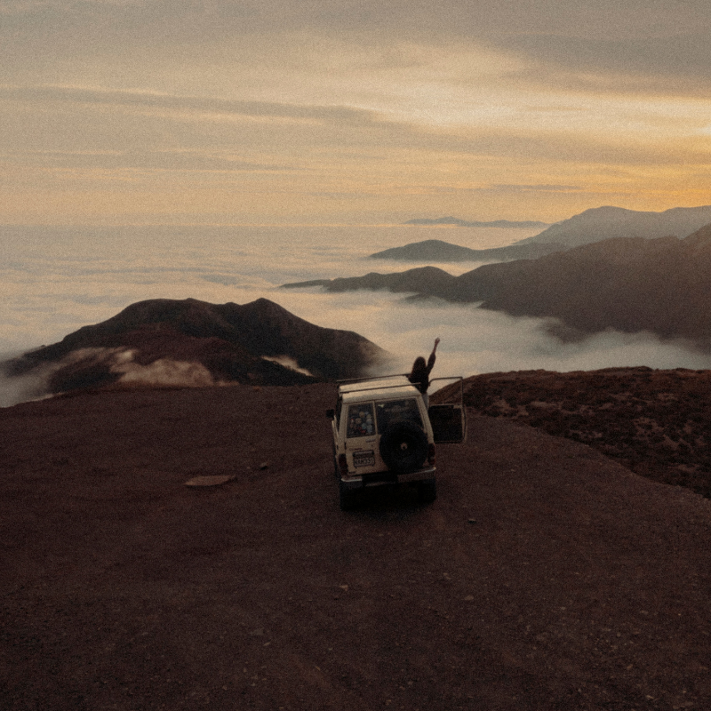 Off-road van parked on a rocky ridge at sunset, with a person standing by the open door and arms raised, overlooking a sea of clouds and distant mountains.