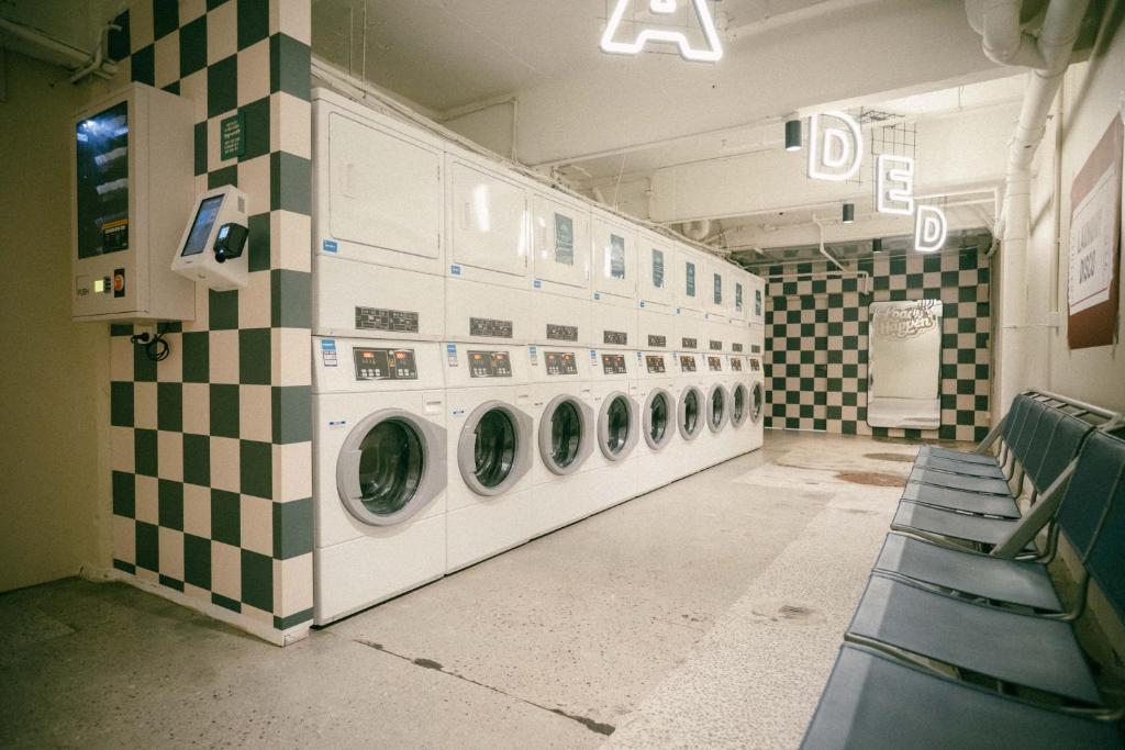 Laundromat with a row of front-loading washers along a checkered green-and-white wall and blue chairs along the right. A long row of coin-operated washing machines in a retro laundromat with neon letters hanging from the ceiling.