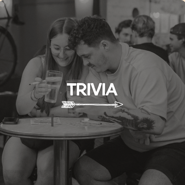 Two friends at a cafe table play trivia, smiling as they look at a phone and notes with a bold 'TRIVIA' overlay.