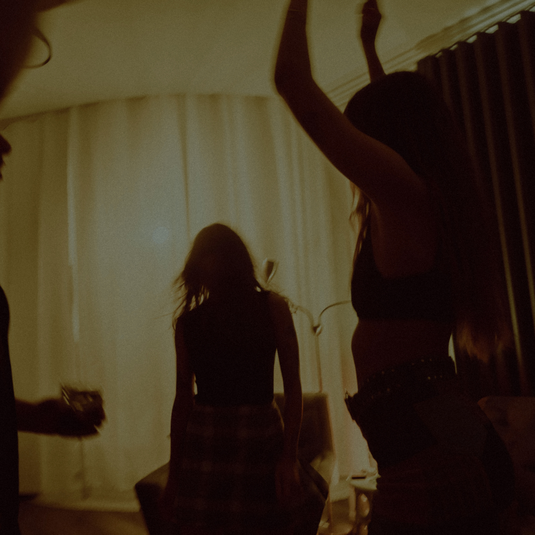Silhouettes of people dancing indoors, with one person raising arms against a warm, dimly lit room backdrop.