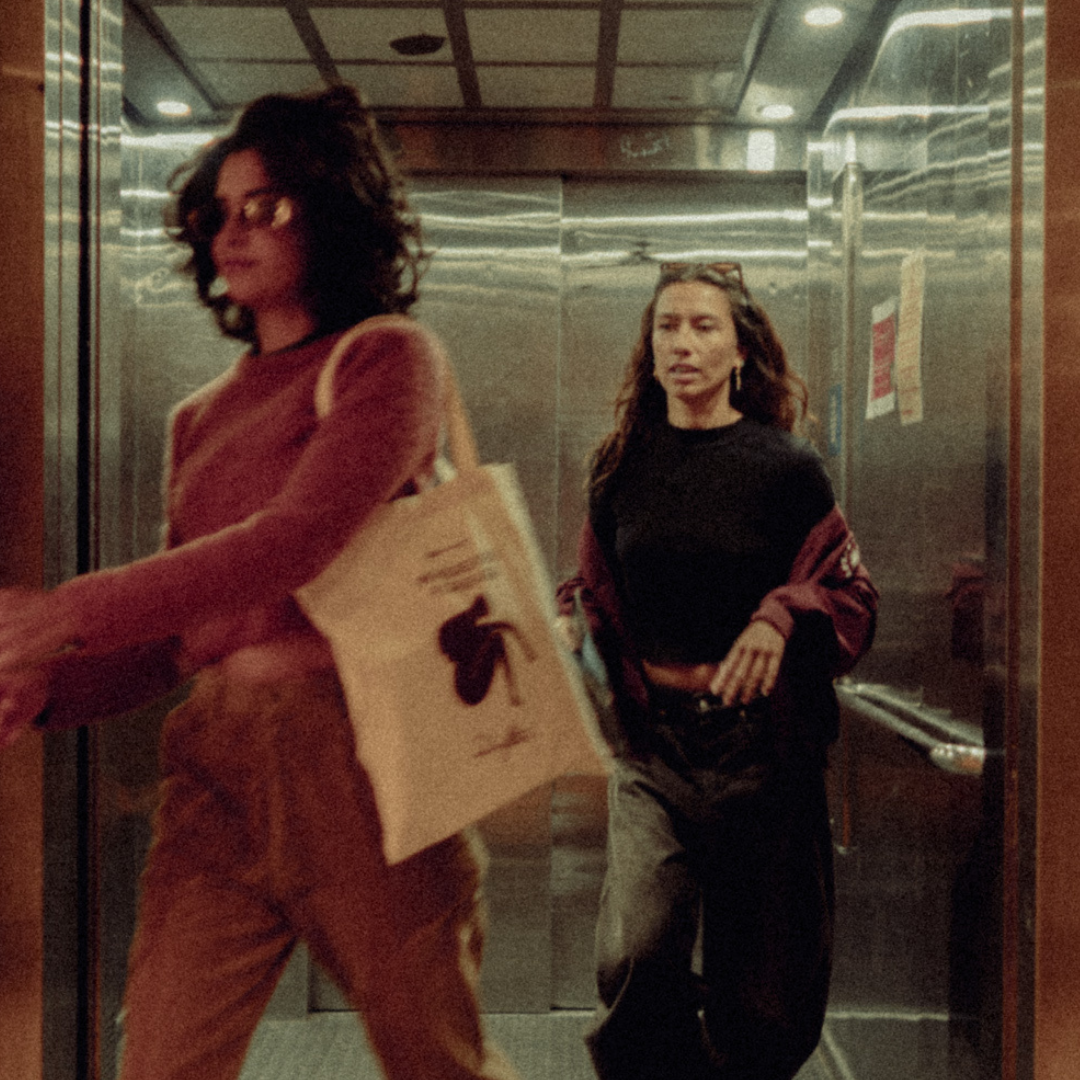 Two women in an elevator; one in an orange outfit carries a tote bag and papers, the other in a black top with a maroon jacket.