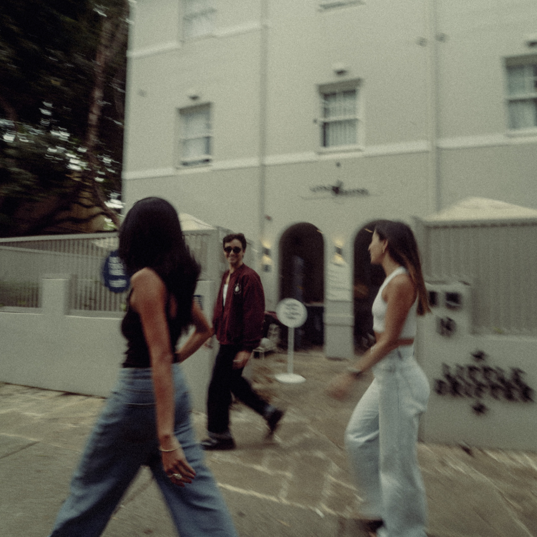 Three people walking along a sidewalk past a pale-green building, with a man in a red jacket between two women (motion blurred).