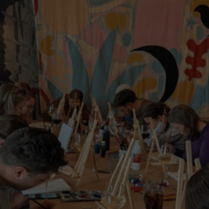 Group of people in an art class painting small wooden easels at a long table, with a colorful mural on the wall behind them.