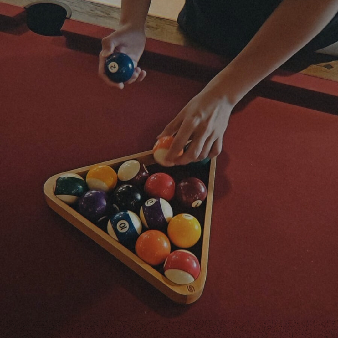 Hands place a pool ball into a triangular rack filled with colorful billiard balls on a red pool table.