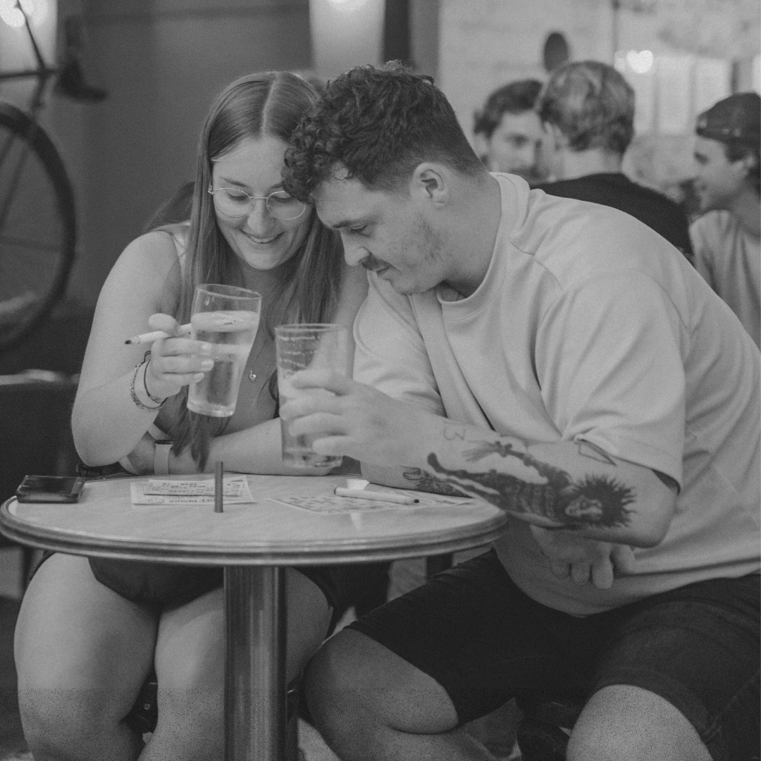 Young couple at a round café table, smiling and sharing drinks while looking at papers on the table.