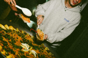 Two people slice and serve a large herb-topped pizza with lemon wedges on a wooden table.
