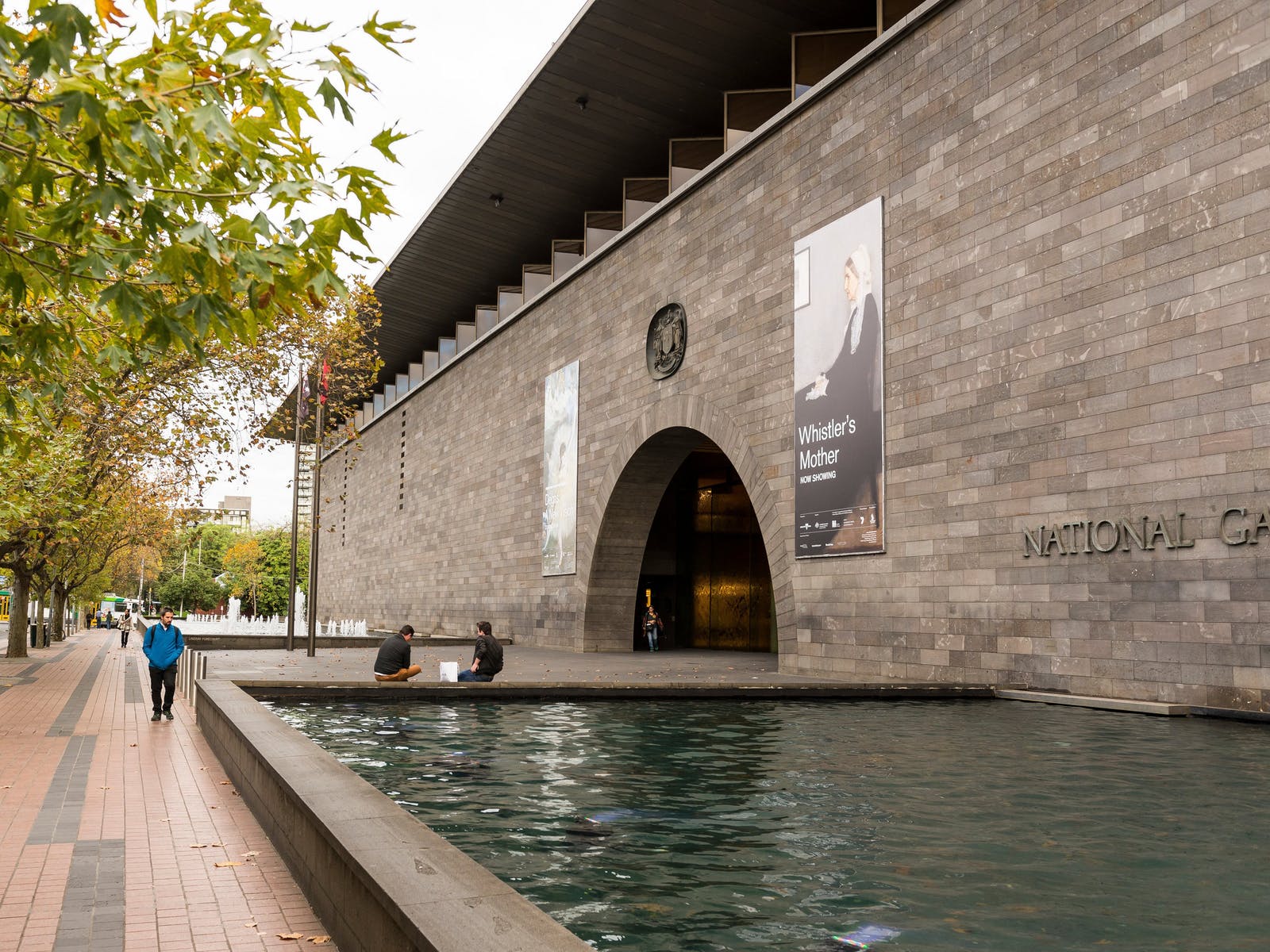 Wide view of a stone National Gallery building with an arched entrance and banners, beside a reflecting pool and trees along a brick walkway.