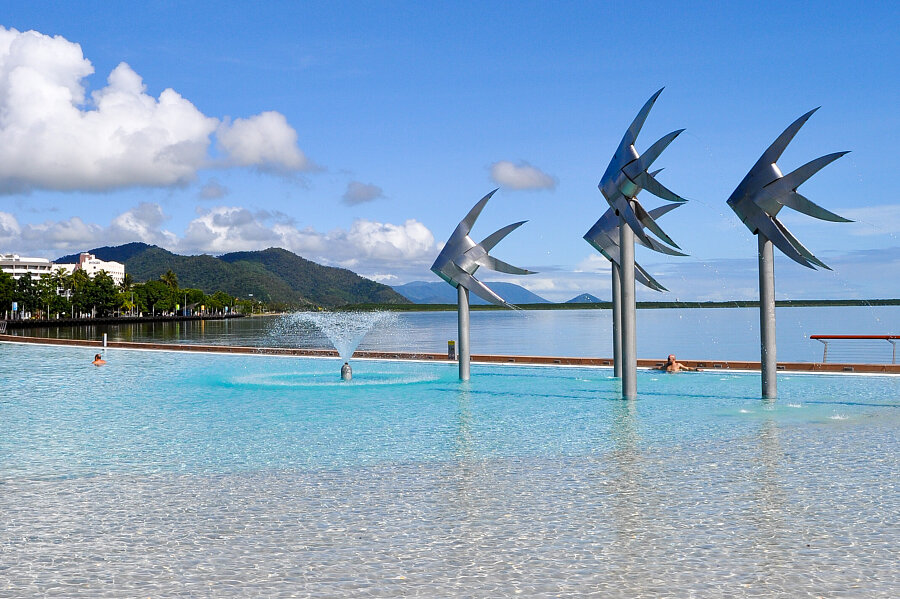 Outdoor pool with four metal bird sculptures on posts, a central fountain, and distant hills under a bright blue sky.