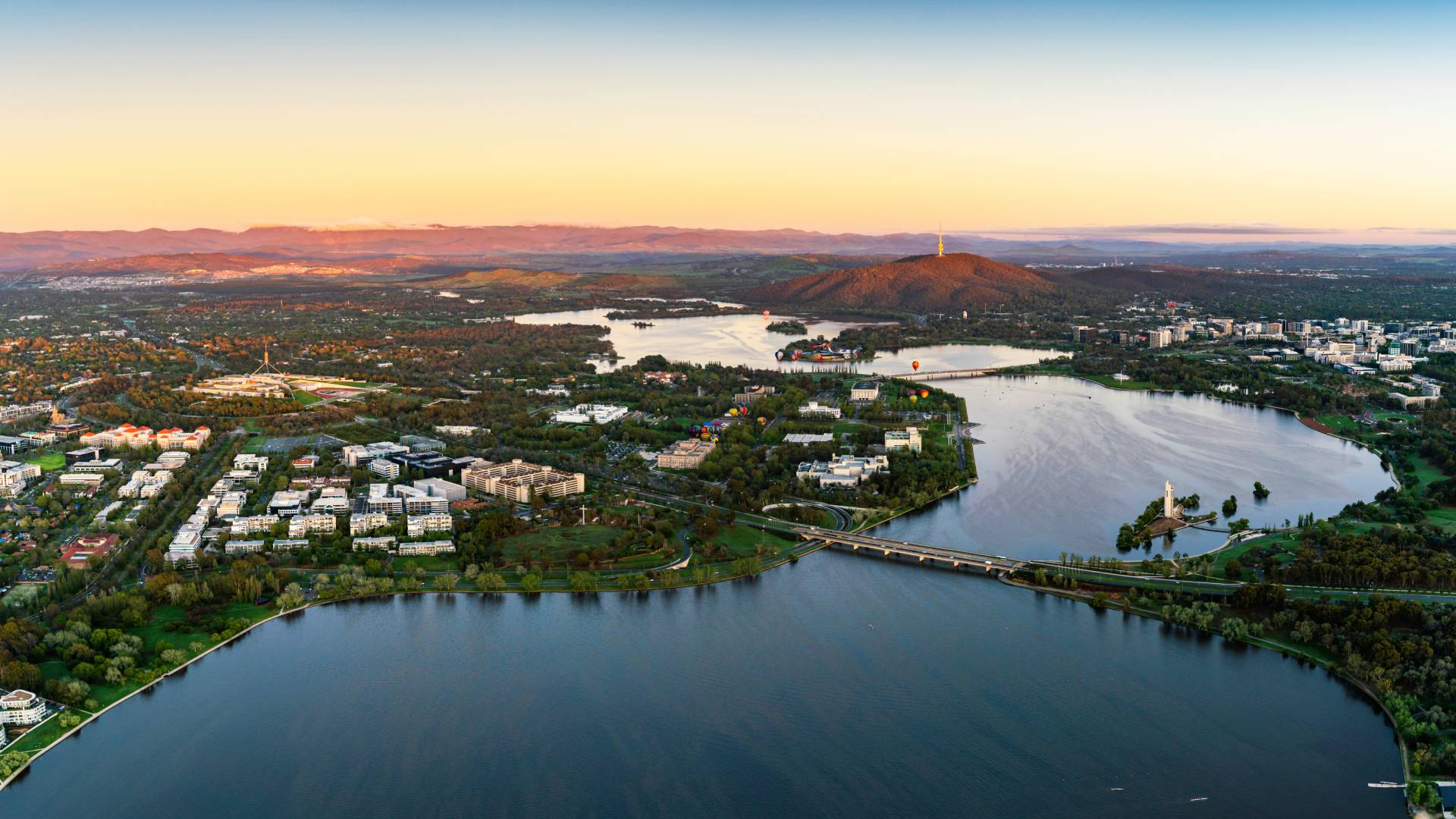 Aerial view of a city along a winding river at sunset, with bridges, green spaces, and a hill crowned by a communications tower in the distance?bed