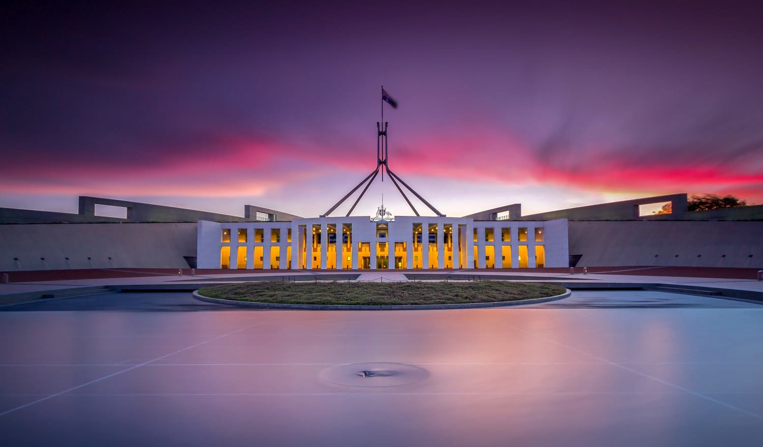 Parliament House in Canberra at sunset, with glowing windows and a circular forecourt.