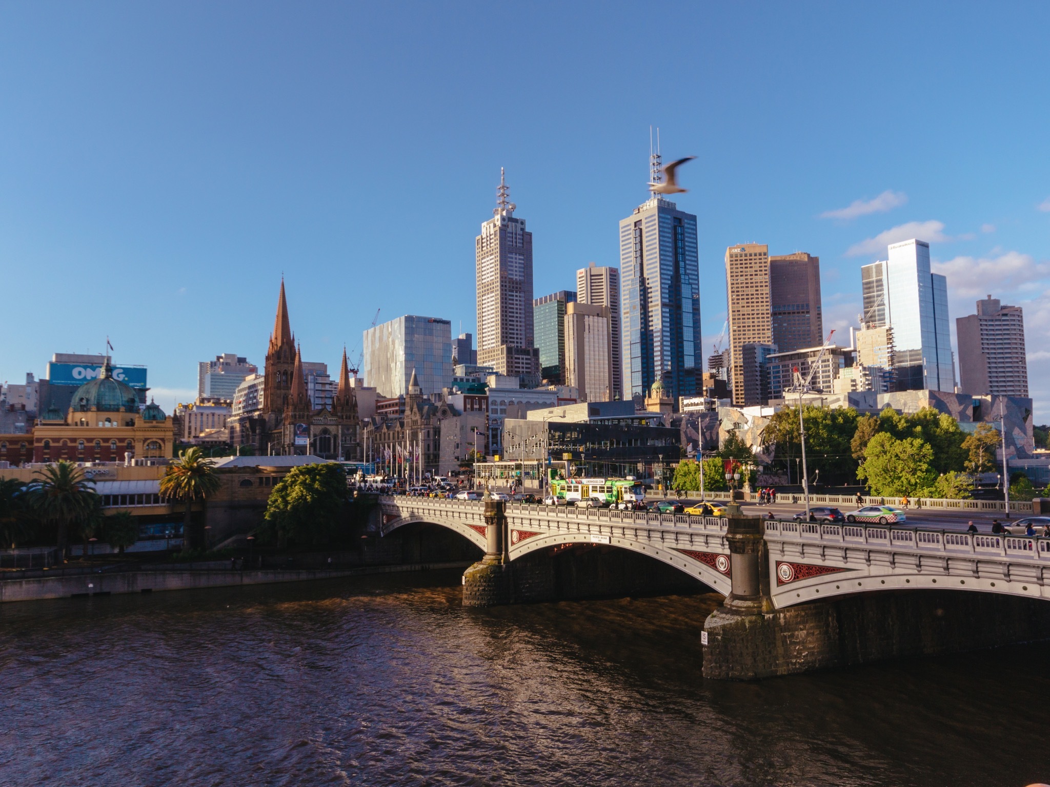 City skyline along a river with a decorative stone bridge in the foreground and modern high-rise buildings against a clear blue sky.