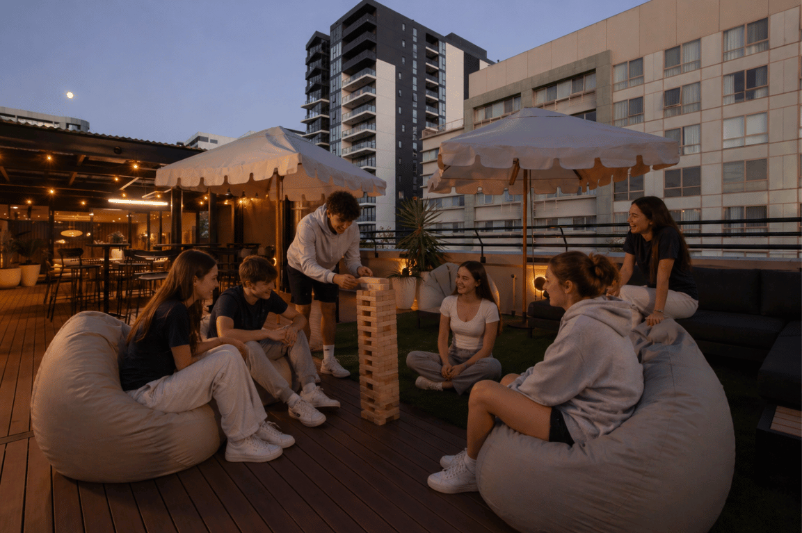 Group of six friends on a rooftop deck playing a wooden block Jenga-style game, seated on beanbags at dusk with city buildings in the background.