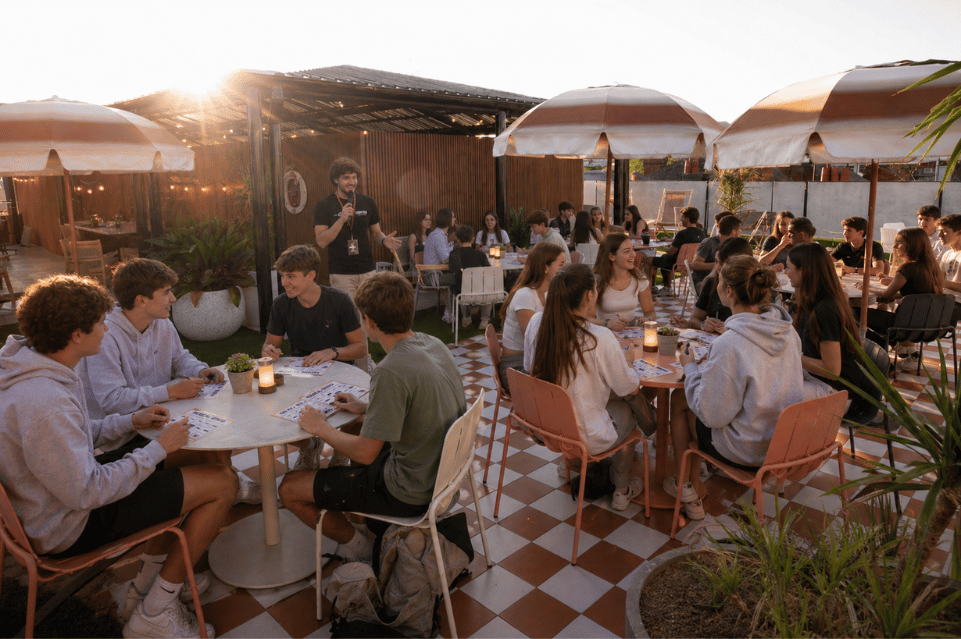 Outdoor patio restaurant with groups of young people chatting and eating around round tables as a man speaks into a microphone under a shaded area, sunset light.