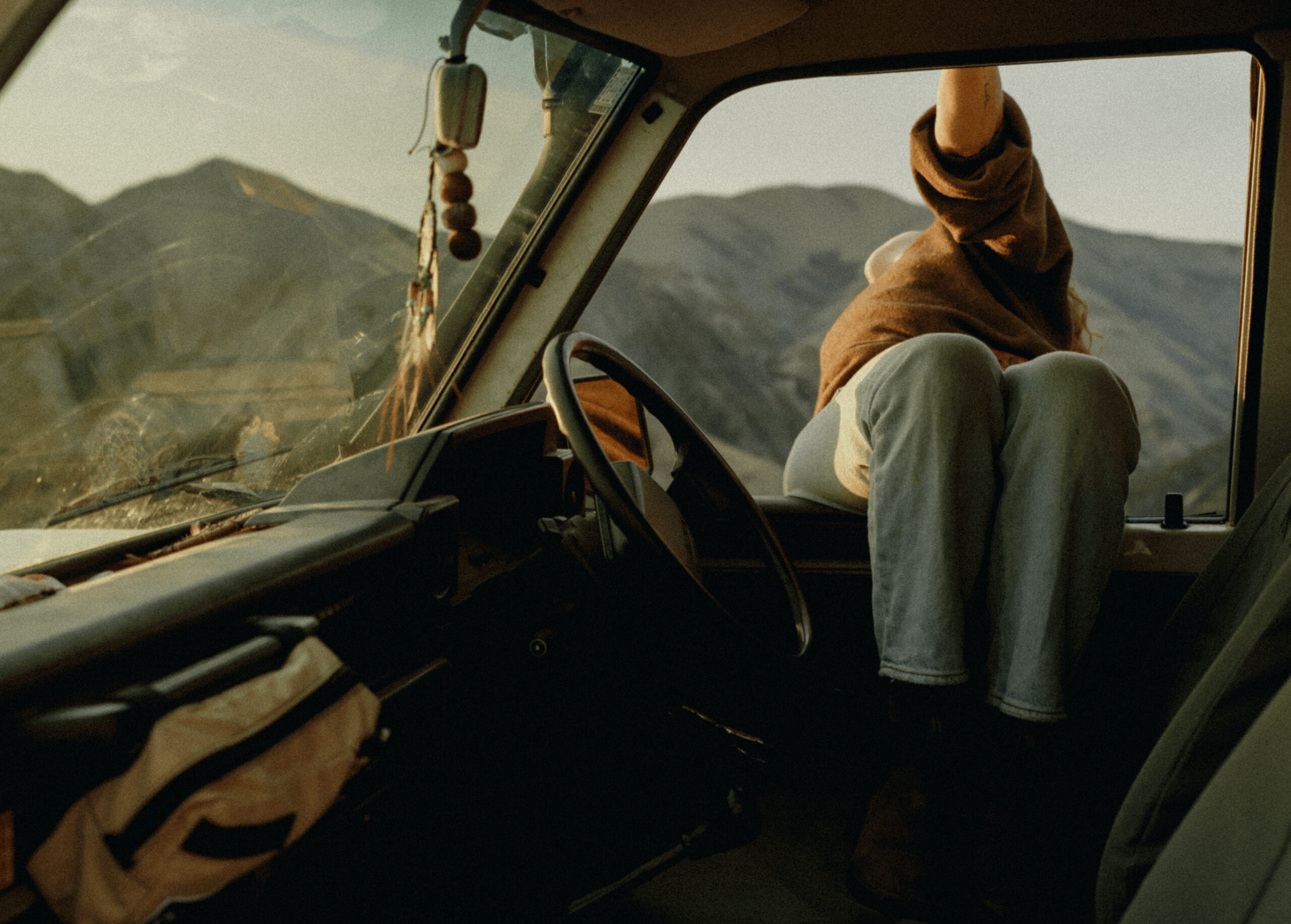 Person in a brown hoodie raises an arm through the vehicle window, with mountains in the background inside the cockpit.