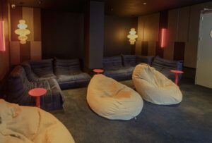 Dimly lit lounge with beige bean bags and blue couches arranged around small pink tables.