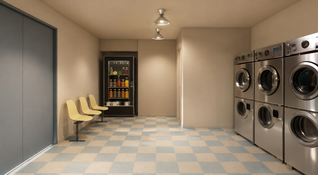 Laundromat hallway with a row of front-loading washers on the right, beige walls, and blue-tan checkered floor; yellow chairs line the left wall facing a vending fridge in the back corner.