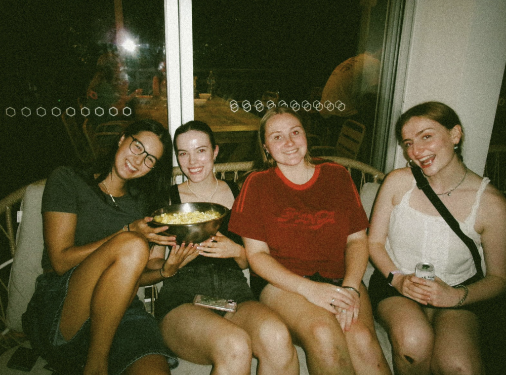 Four friends sit together on a couch at night, smiling; one holds a bowl of popcorn in her lap.