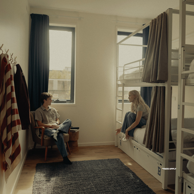 Two people converse in a dorm room: a man sits in a chair while a woman sits on a lower bunk, with a bunk bed, striped towel, and two large windows nearby.