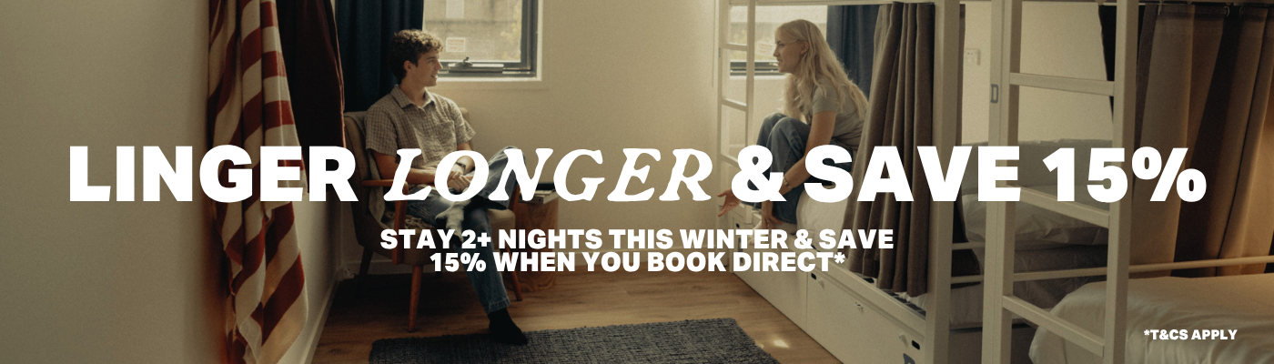 Two people sit in a dorm-style room with bunk beds; bold overlay text reads 'LINGER LONGER & SAVE 15%' for a winter promotion.
