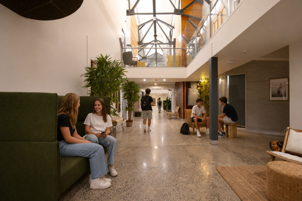 Two young women sit on a green sofa in a modern, plant-filled lobby and chat as others lounge nearby.