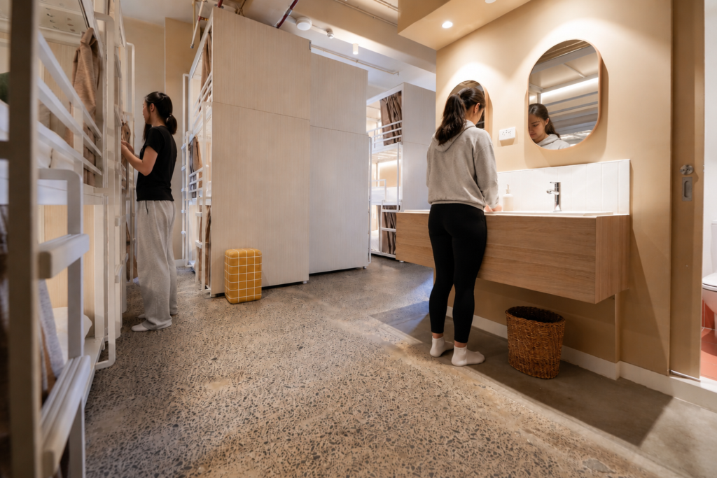 Woman at a white-tiled bathroom sink in a warm dorm-style restroom with wooden vanity and round mirror.