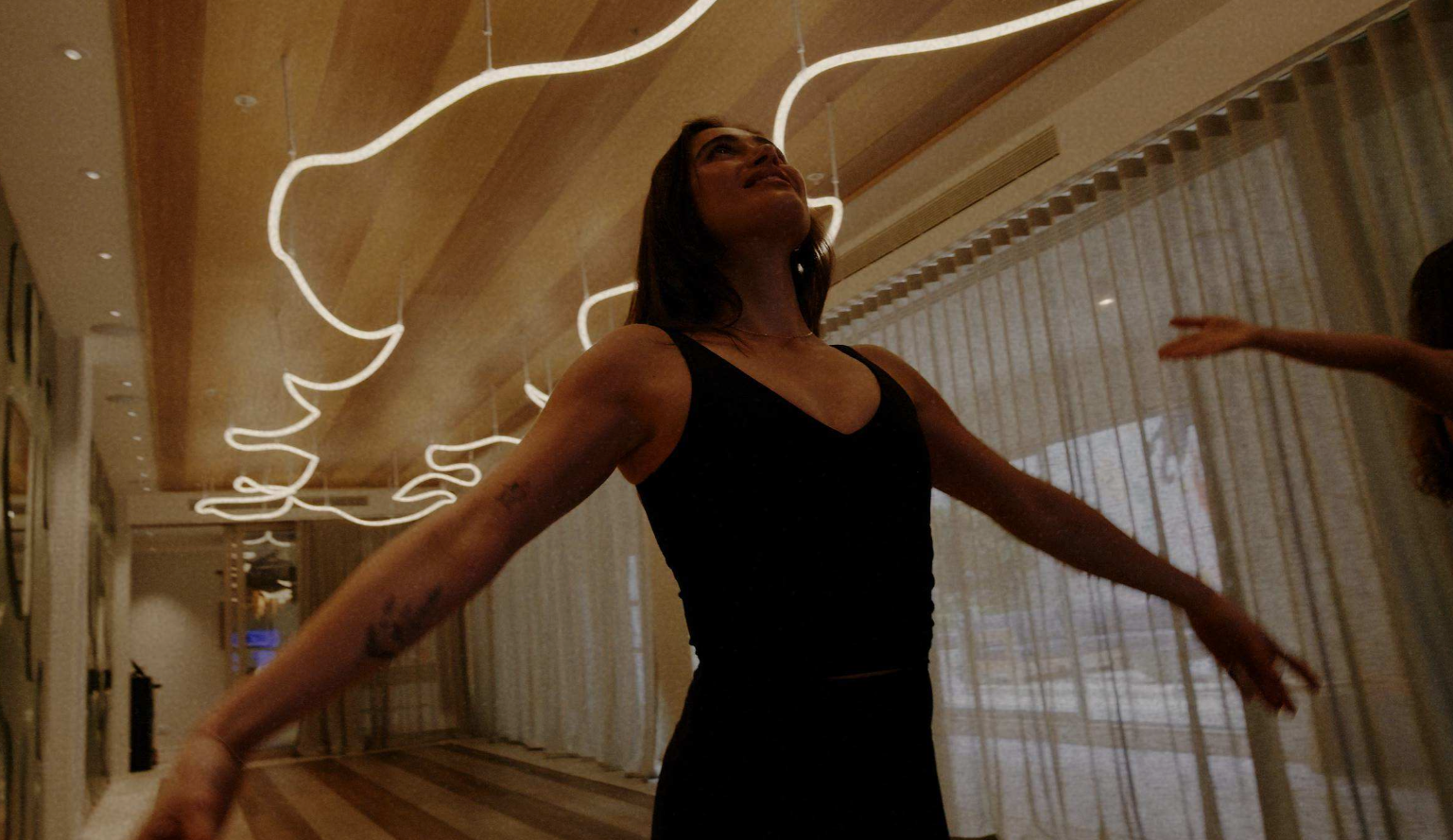 Woman in a black sleeveless dress dancing with arms outstretched in a hallway lit by abstract neon lights overhead.