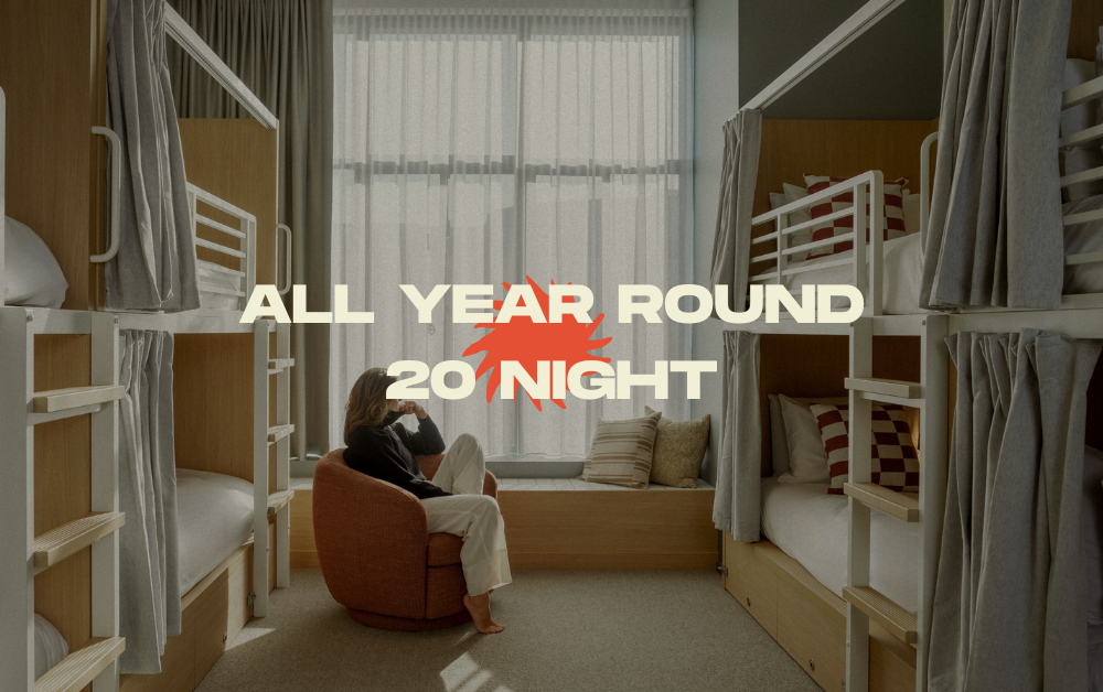 Promotional banner in a hostel dorm room with a person sitting in an orange chair; overlay text reads 'ALL YEAR ROUND 20 NIGHT'.