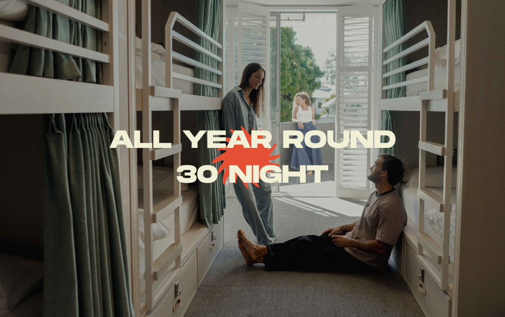 Dorm-style hostel room with bunk beds; two people stand near the doorway, promo text 'ALL YEAR ROUND 30 NIGHT' across the center.