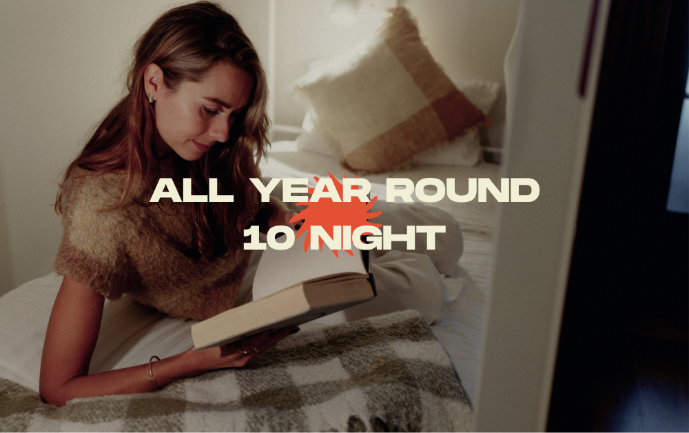 Woman reading a book on a cozy bed in a softly lit bedroom, with the words ALL YEAR ROUND NIGHT across the image.