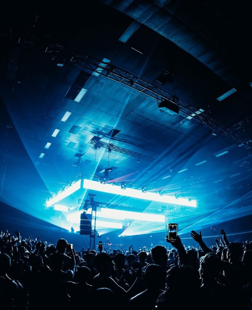 Crowd at a blue-lit concert with raised hands and phones, a bright stage and lasers in the background.