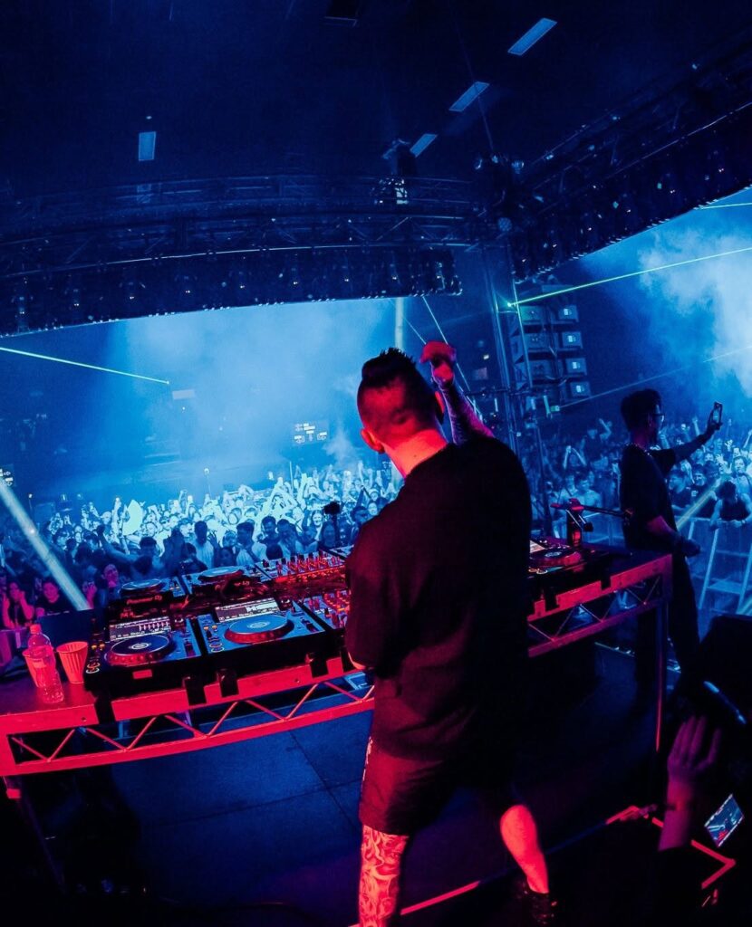 DJ at a mixer and turntables on a crowded club stage, blue and pink lights, and a cheering audience in the haze.