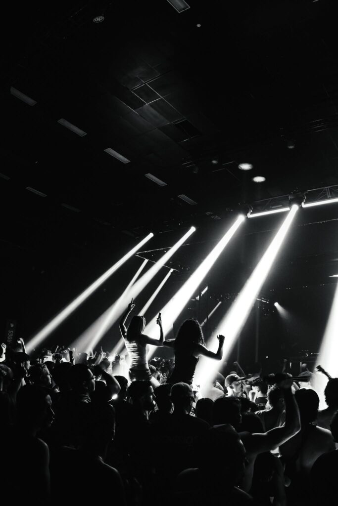 Silhouetted concert crowd with raised arms beneath bright diagonal white stage lights in a dark venue.