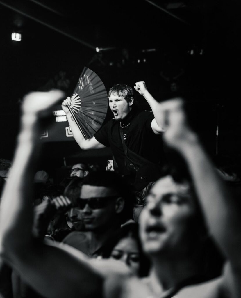 Young man on a crowd-surfed barrier raises a fist and holds a fan, cheering at a concert.