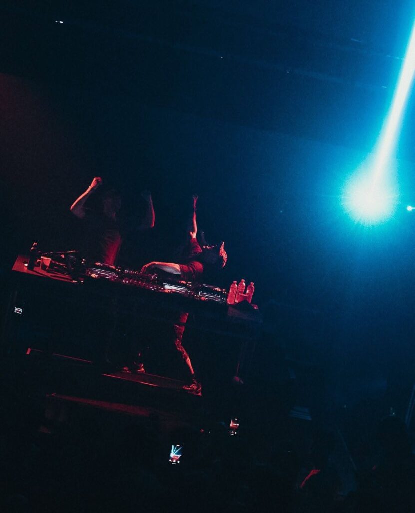 DJ performing on a dark stage with equipment and bottles, red lighting, blue spotlight on the right, crowd raising hands
