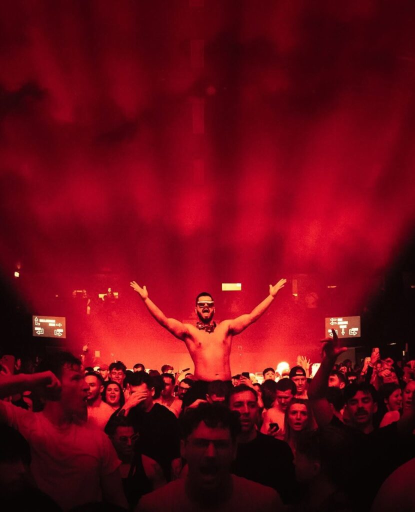 Shirtless man with arms raised stands on someone's shoulders in a crowded club under red stage lights and fog.