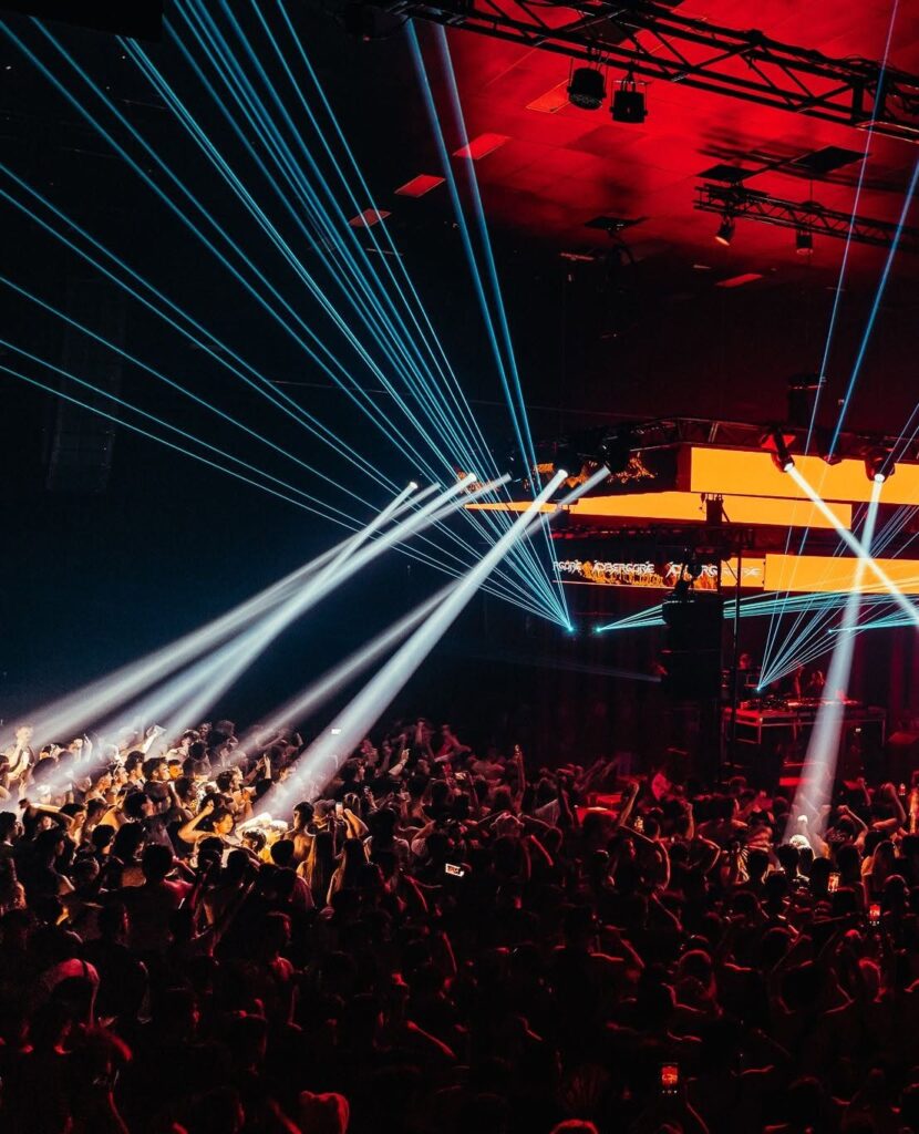 Crowd at a nightclub with bright blue laser beams crossing above a stage with orange screens and red lighting