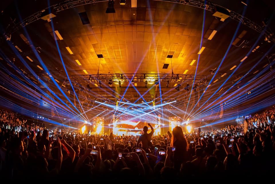 Wide shot of a large concert crowd facing a brightly lit stage with orange and blue laser lights streaking across the arena.