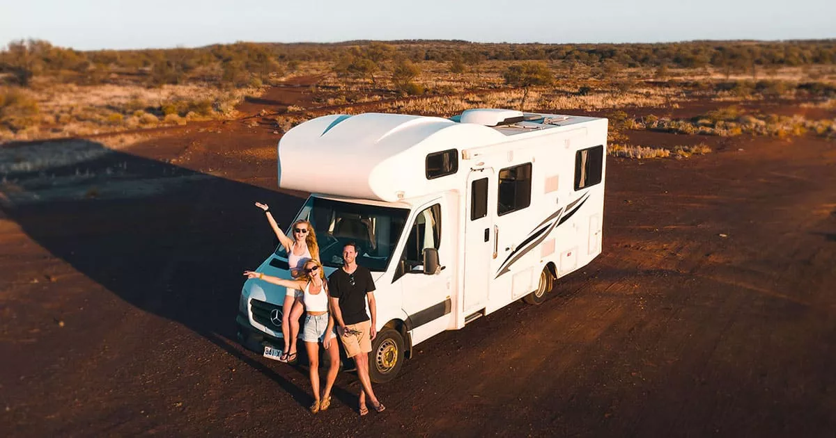 Four friends posing by a white camper van in a dry desert landscape at sunset.