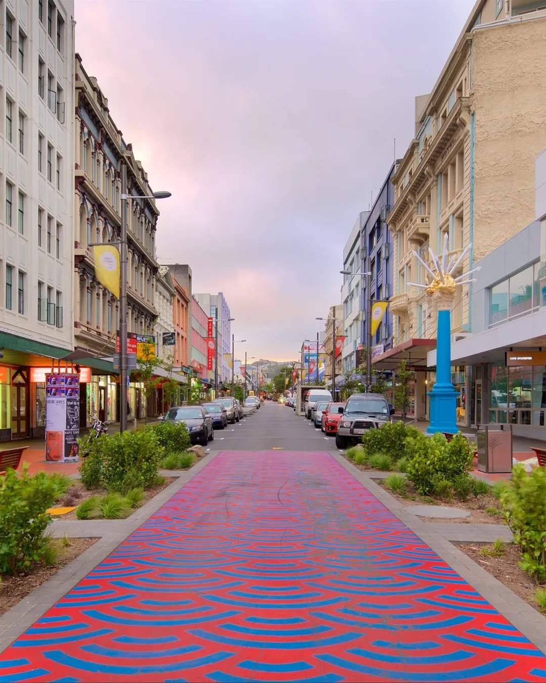 A vibrant city street with a red-and-blue patterned pedestrian walkway, pastel buildings, parked cars, and a bright blue sculpture pillar on the right.