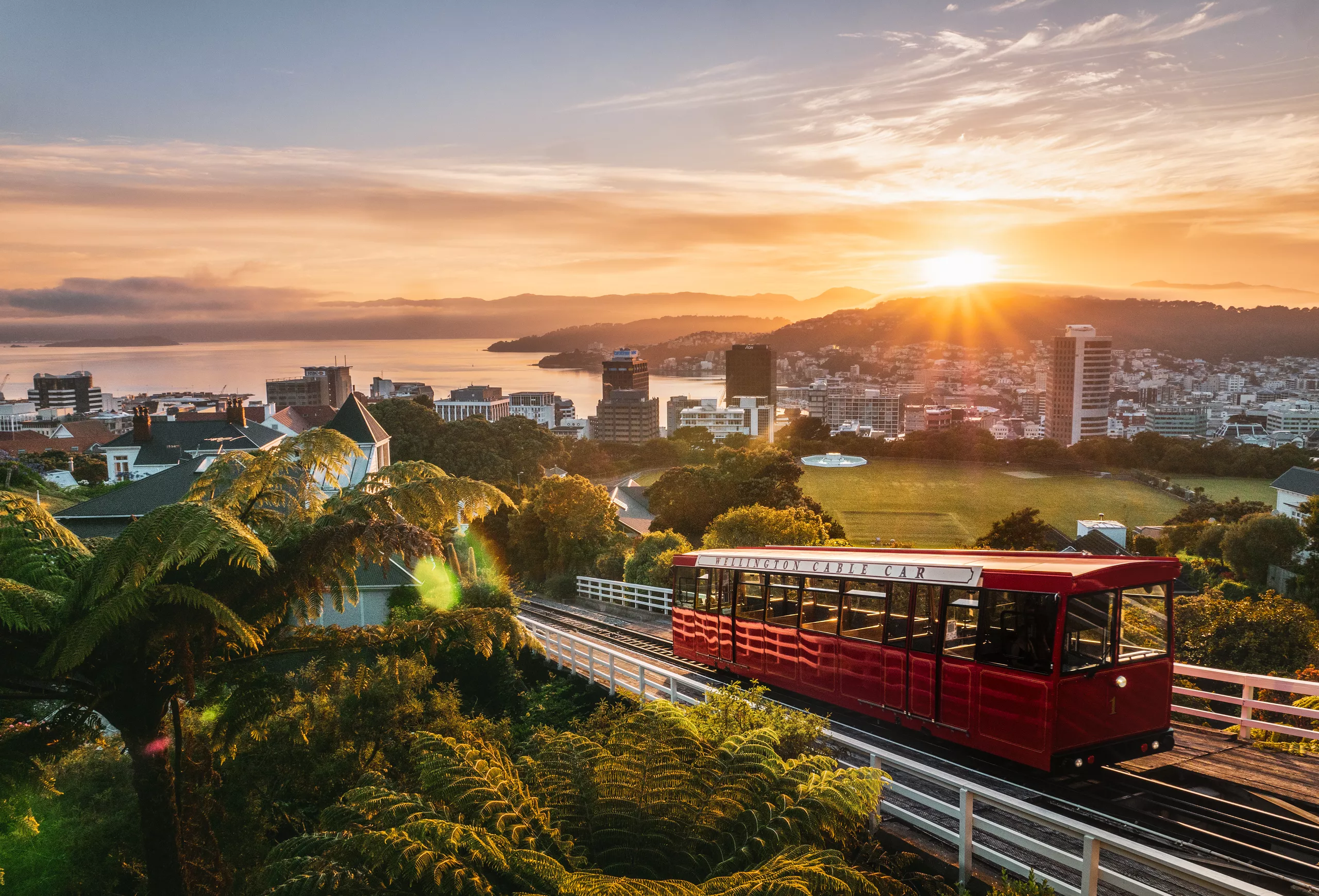 Sunset over Wellington city with a red cable car on tracks in the foreground and water beyond the hills.