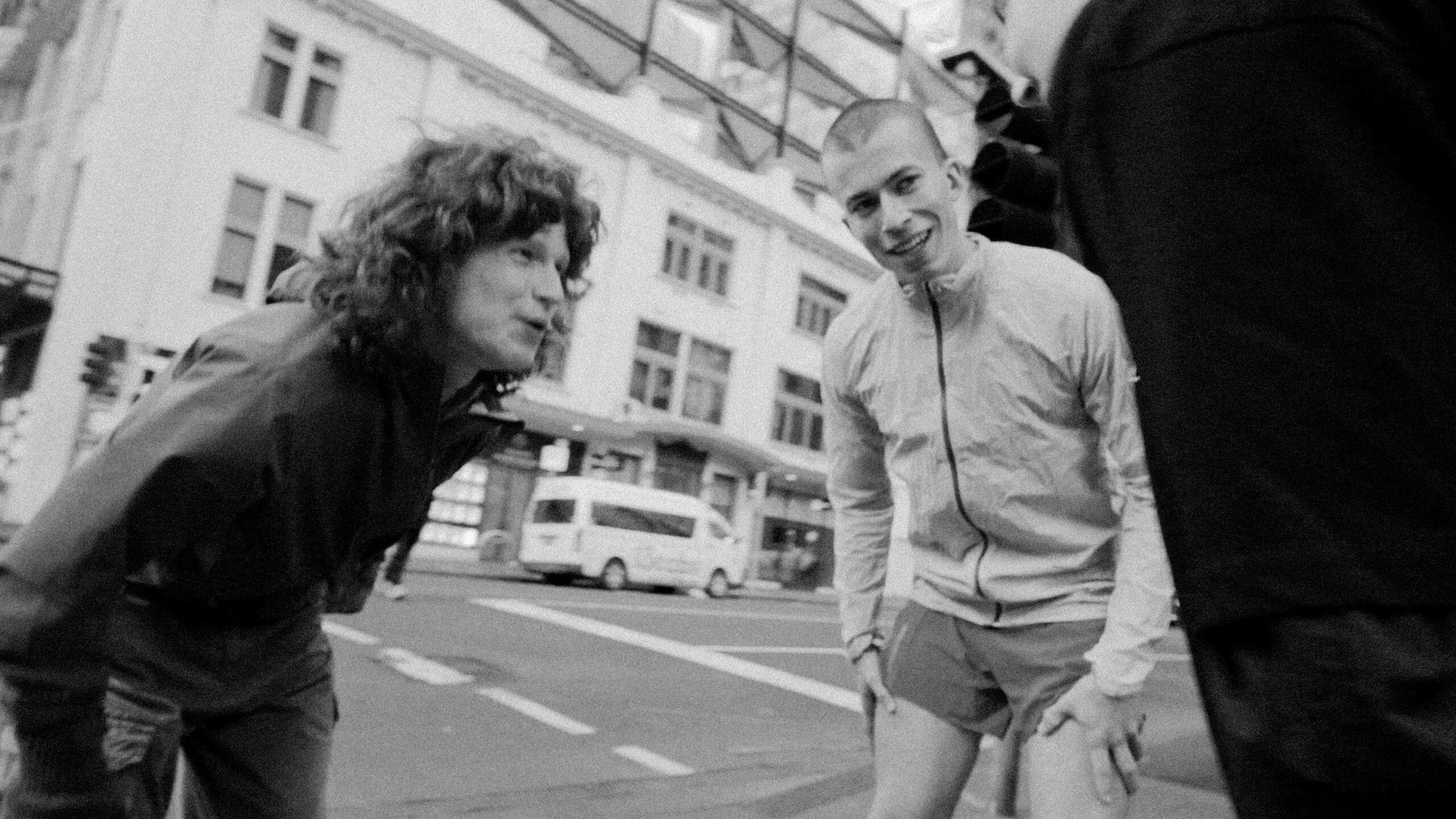 Two people leaning toward the camera and smiling on a city street, with a bus and buildings in the background (black-and-white photo).