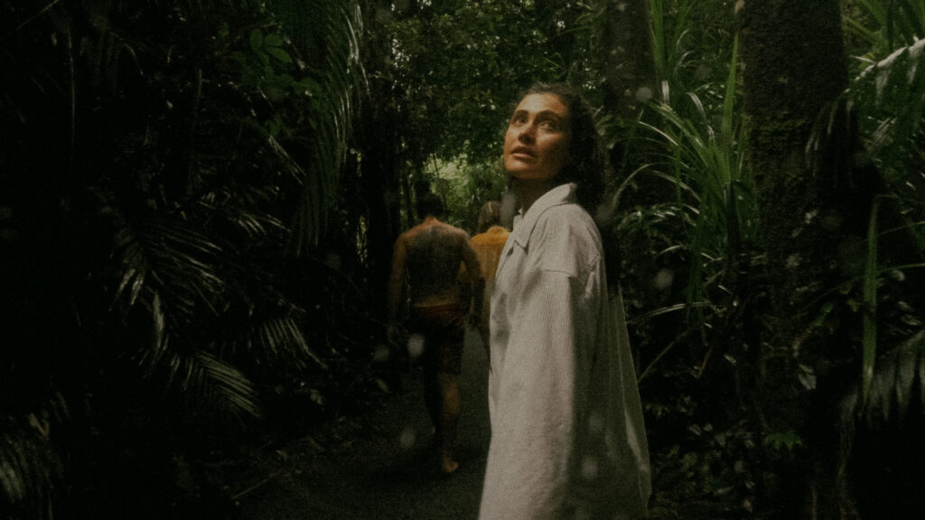 Woman in a light-colored dress standing on a shaded jungle path, gazing upward among dense tropical foliage, with rain-forest greenery surrounding her