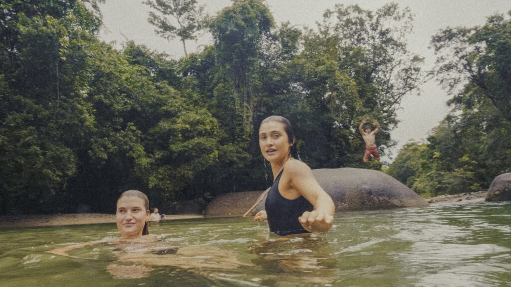 Two women swimming in a river with a forested bank in the background; one woman in a black swimsuit is close to the camera, another behind smiling in the water.