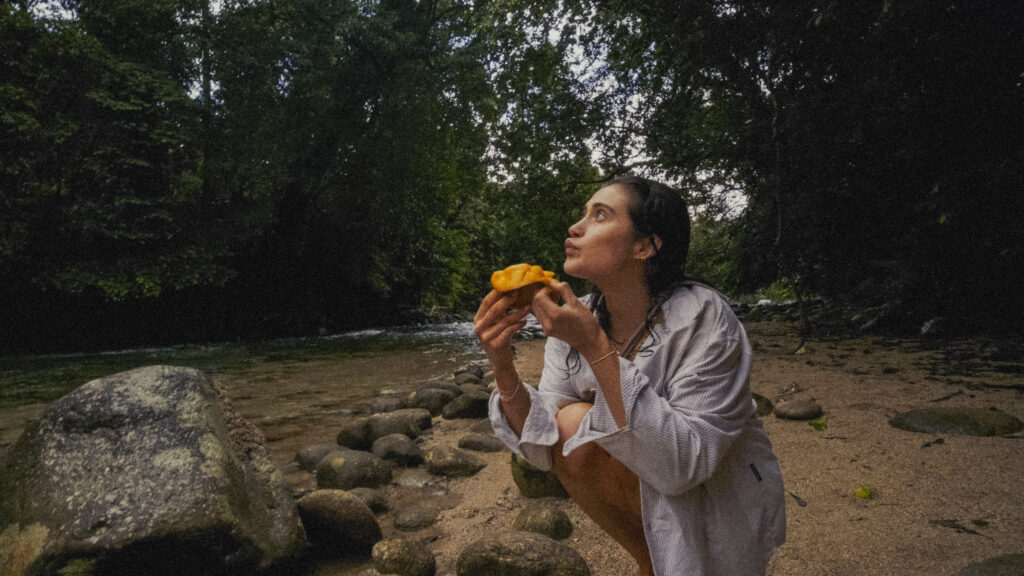 Person crouching on a rocky beach by a stream, wearing a striped shirt and holding an orange cloth near mouth as if eating or smelling it, with trees in the background.