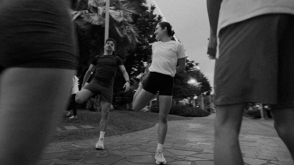 Black-and-white photo of a group walking outdoors on a brick path; center young woman in a white shirt and black shorts smiles as others pass by.