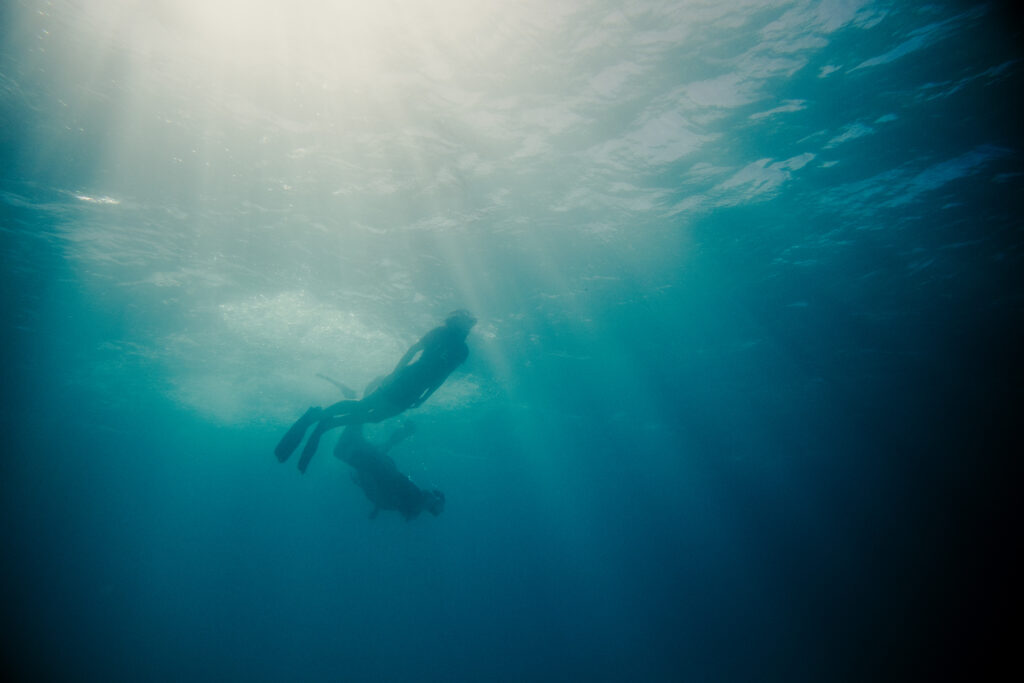 Two scuba divers swimming underwater as sunlight beams through the blue water.