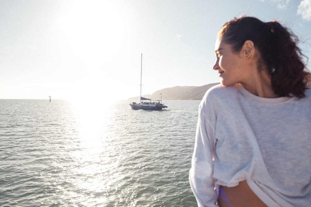 Woman standing near the water, looking toward the horizon as a sailboat sits on the calm sea with sunlight glare washing over the scene.