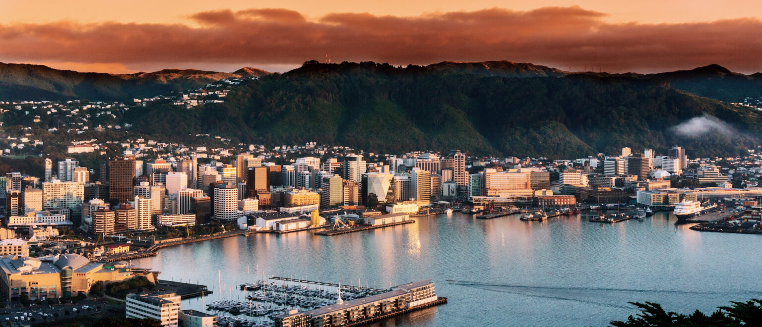 Sunset over a coastal city with a harbor, high-rise buildings, and forested hills in the background.