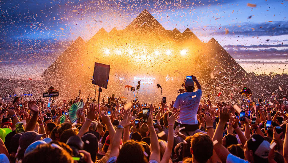 Massive outdoor concert with a pyramid-lit stage, confetti, and a cheering crowd at sunset.